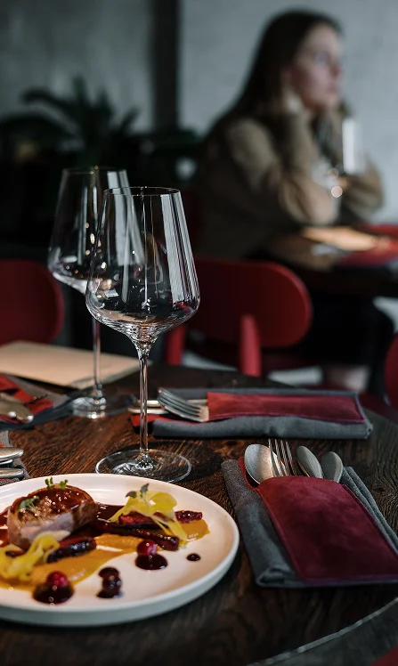 Plat gastronomique dressé sur une table de restaurant sombre, à côté de verres à vin, avec une personne assise à l'arrière-plan.